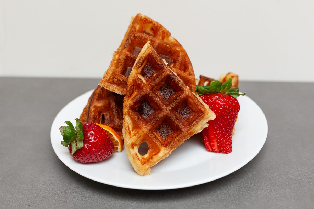 Triangular waffle pieces leaning in a stack on a white plate with whole and halved strawberries and a bit of orange, shot straight