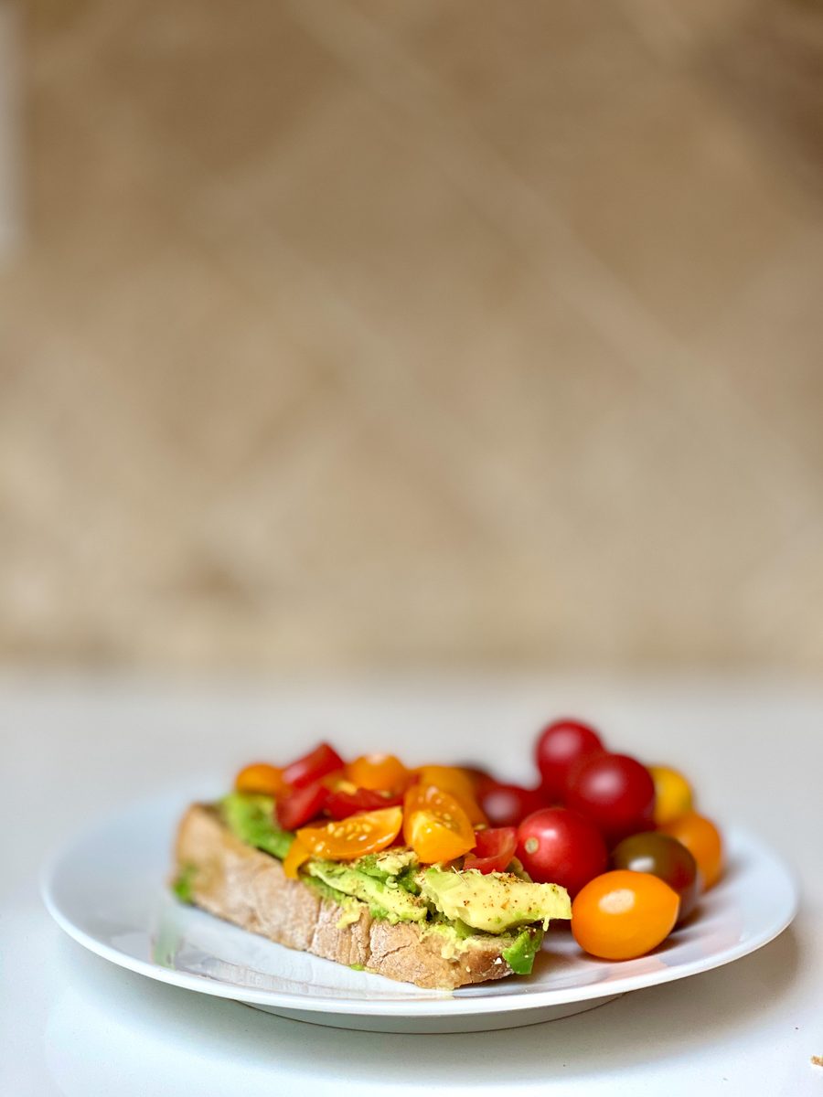 An open-faced avocado toast piled with halved heirloom cherry tomatoes on a small white plate at the bottom of the frame, with a l