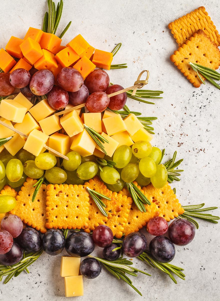Overhead shot of cheese cubes, red and green grapes, crackers and rosemary sprigs arranged into a Christmas tree shape on a white 