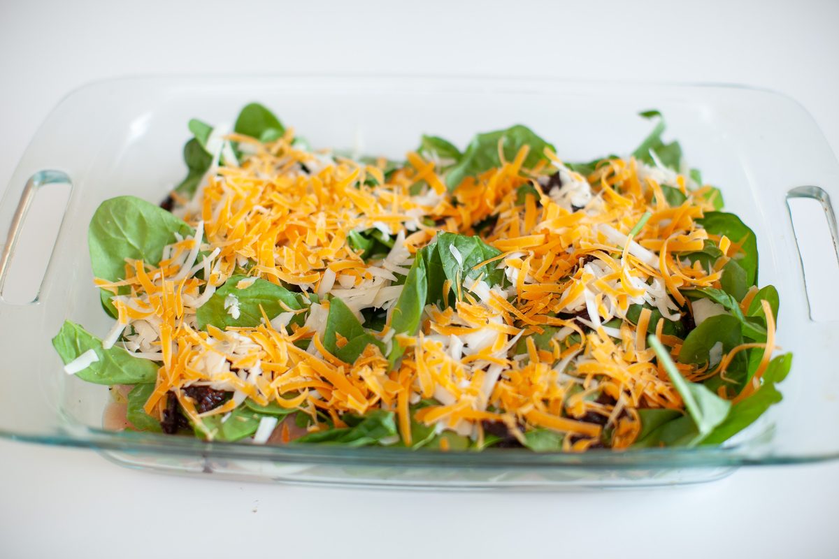 A clear glass baking dish on a white surface filled with fresh spinach leaves under a heavy layer of shredded orange and white che