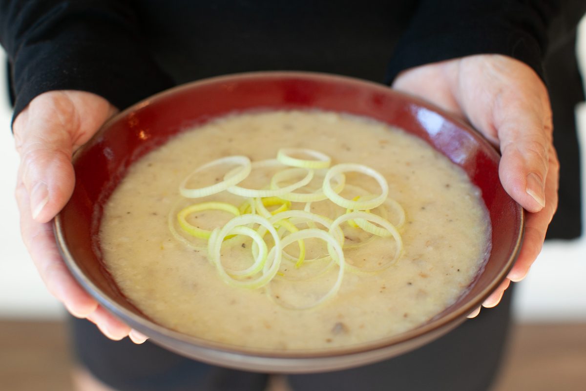Two hands hold out a red-rimmed bowl of thick pale creamy soup garnished with thin rings of leek.