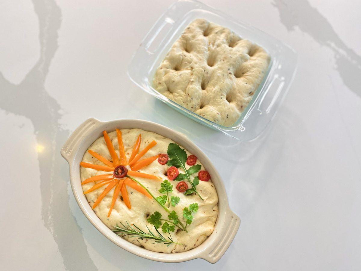 An oval ceramic dish of hummus decorated with a flower pattern of carrot slices, tomato pieces, olives and herb sprigs, with a cov