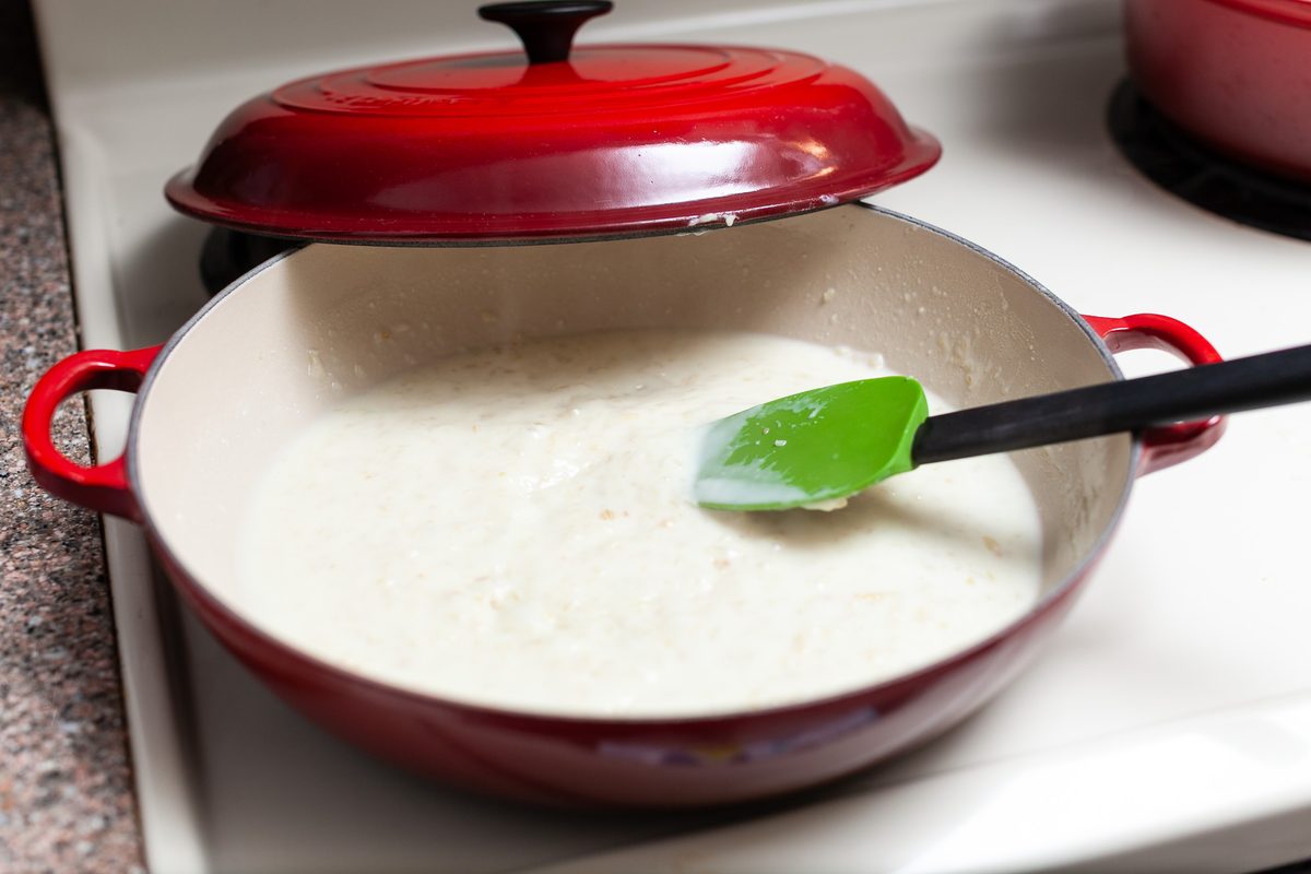A red enameled Dutch oven on a white stovetop filled with thick pale cheese sauce being stirred with a green silicone spatula, lid