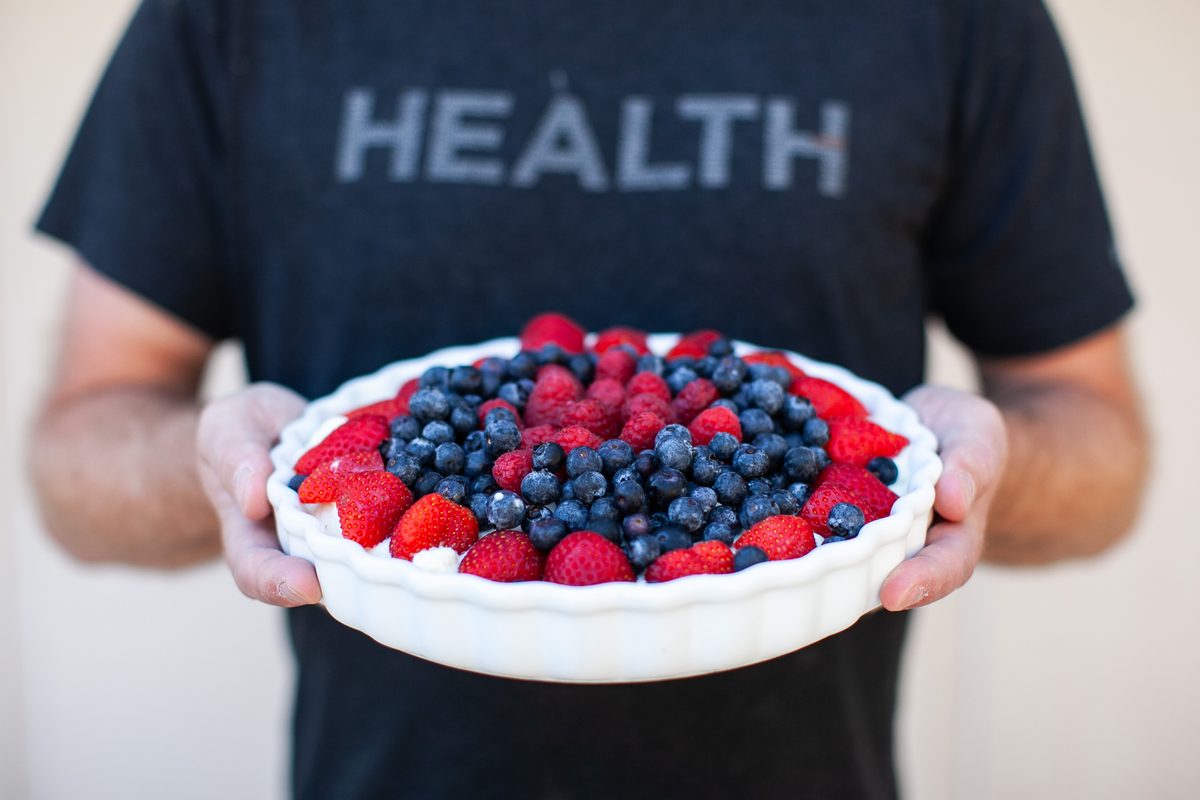 A man in a navy t-shirt printed with the word HEALTH (face out of frame) holding a white fluted dish heaped with fresh strawberrie