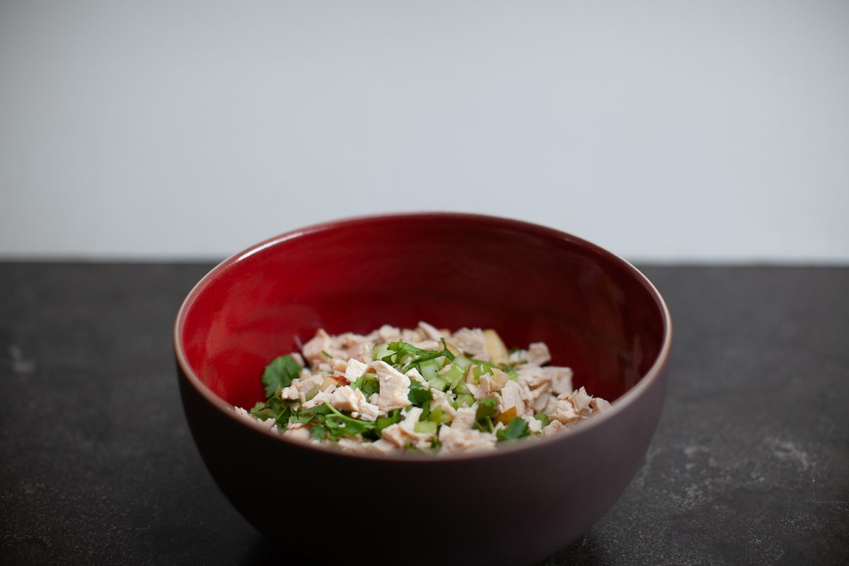 The same dark red bowl of chopped chicken and apple salad with herbs on a dark textured surface, shot dimly against a grey backgro