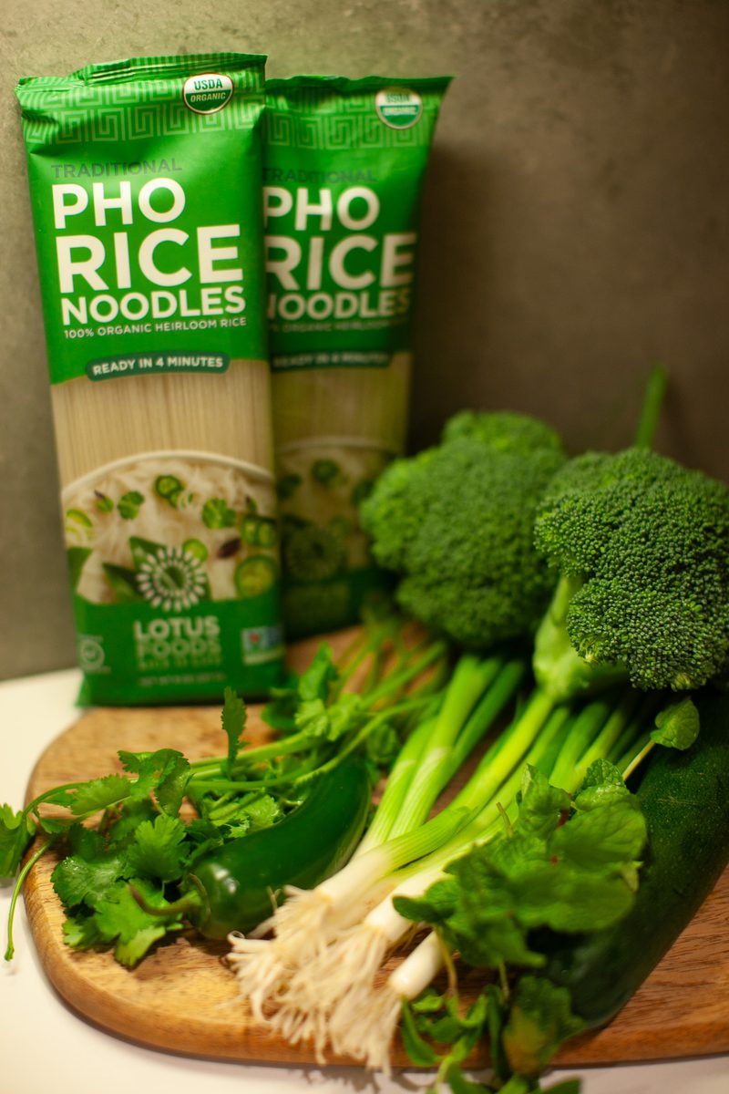 Two sealed green packages of pho rice noodles propped on a wooden board with fresh broccolini, herbs and green onions.