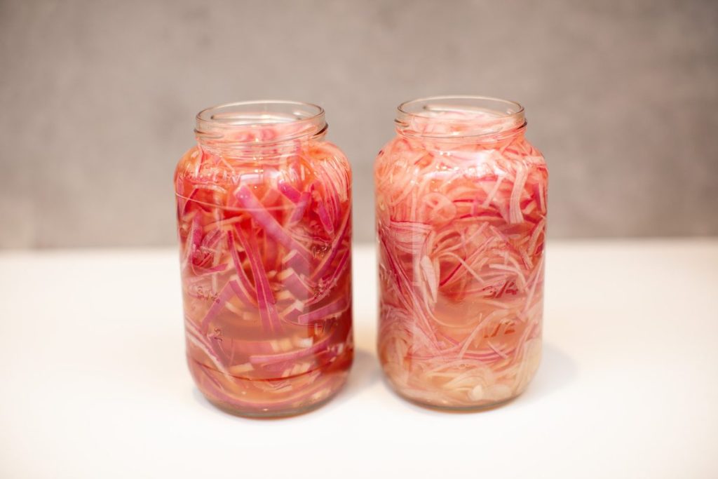 Two glass mason jars packed with bright pink pickled red onions in brine on a plain white surface, no hands.