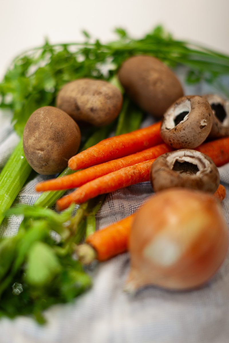 A pile of raw produce on a cloth: whole potatoes, carrots with their tops and other fresh vegetables