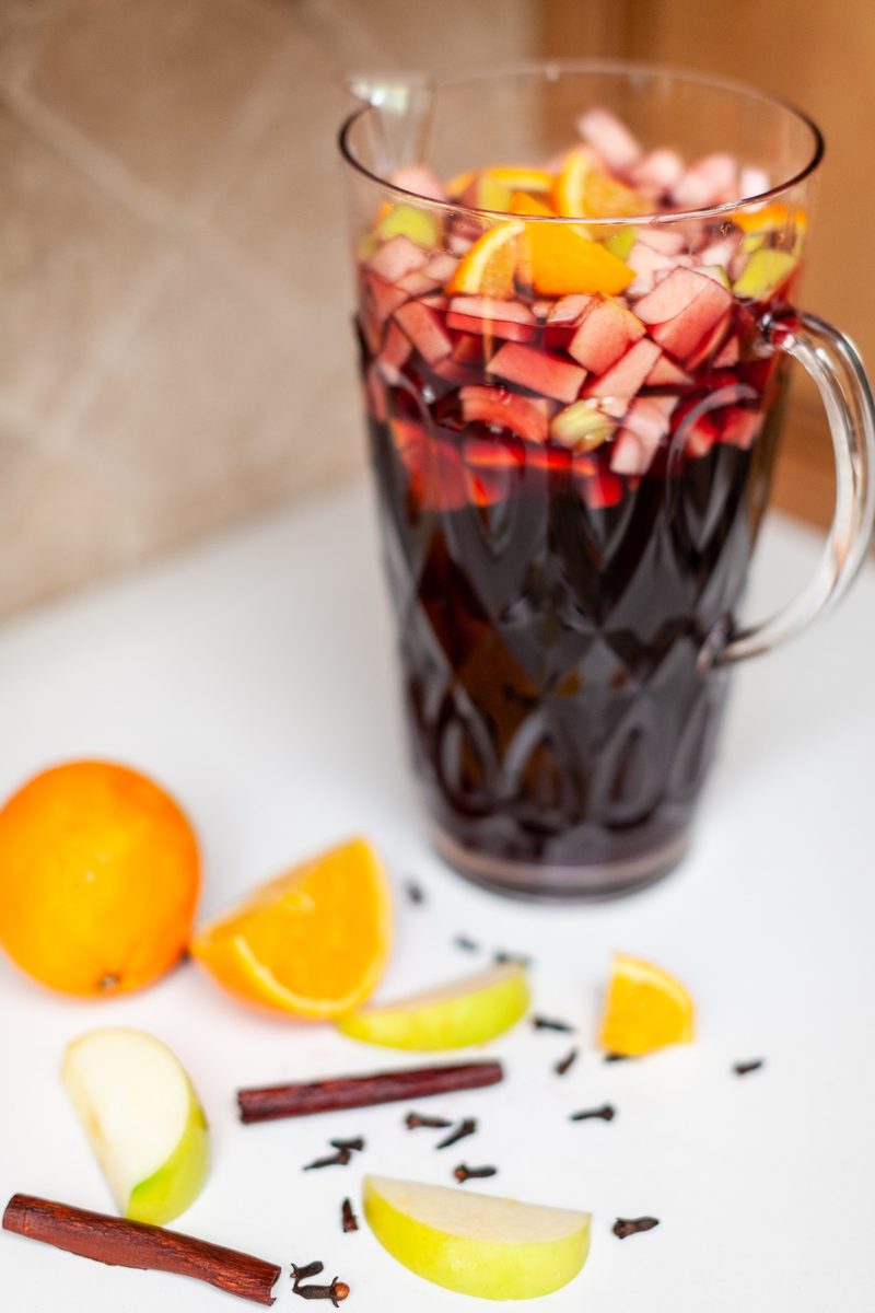 A tall glass pitcher of deep red sangria packed with chopped apple and orange, standing on a white surface with orange halves, app
